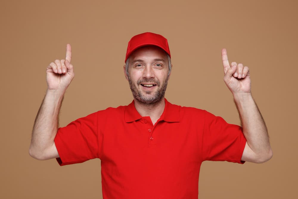 delivery man employee red cap blank tshirt uniform pointing with index fingers up smiling confident looking camera standing brown background Manitas a domicilio Madrid